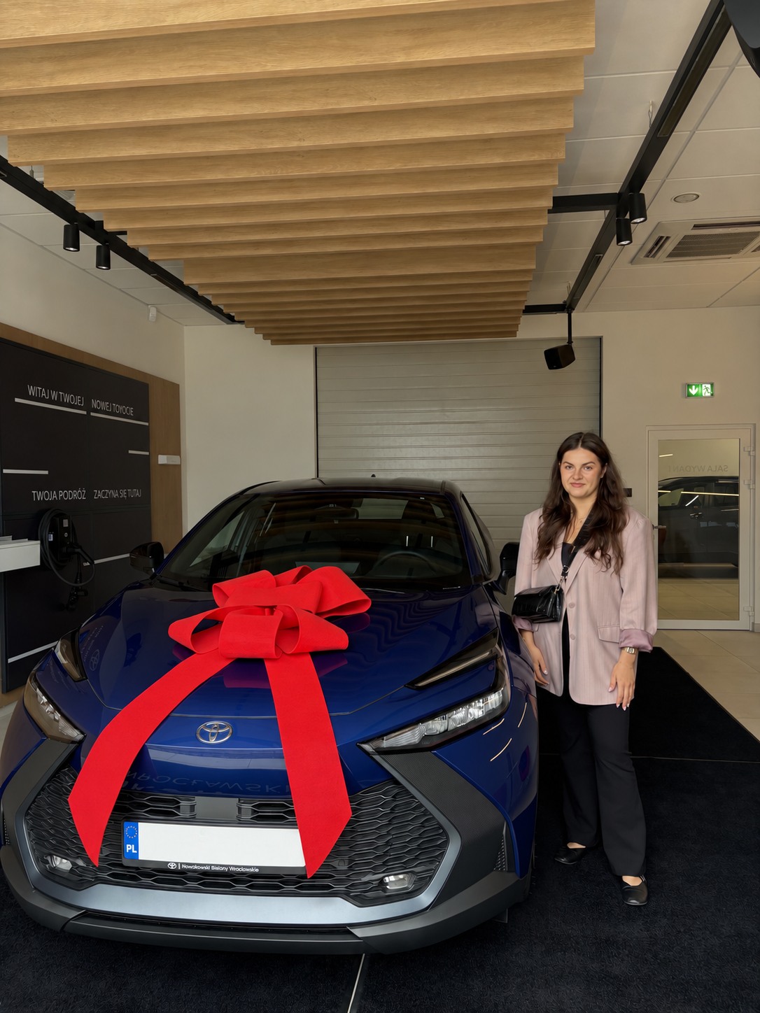Granatowa Toyota z czerwoną kokardą i kobietą stojącą obok w salonie