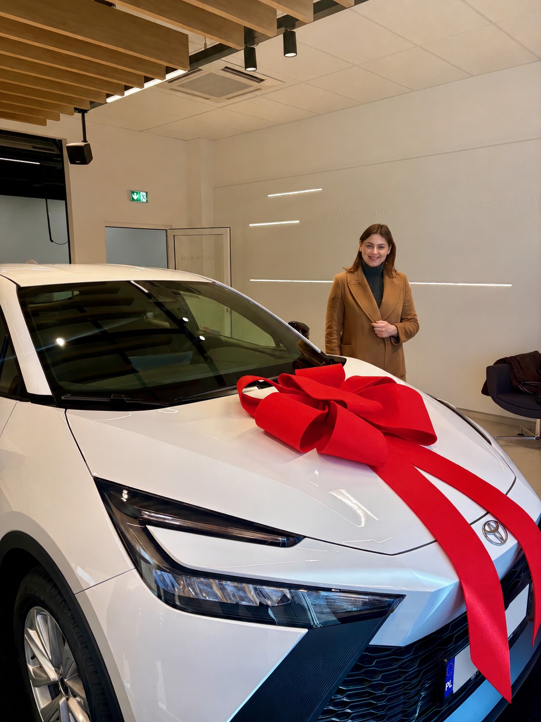 Biała Toyota z czerwoną kokardą i kobietą w brązowym płaszczu w salonie