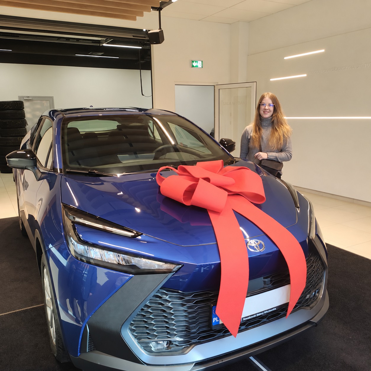 Granatowa Toyota z czerwoną kokardą, obok kobieta w okularach w salonie